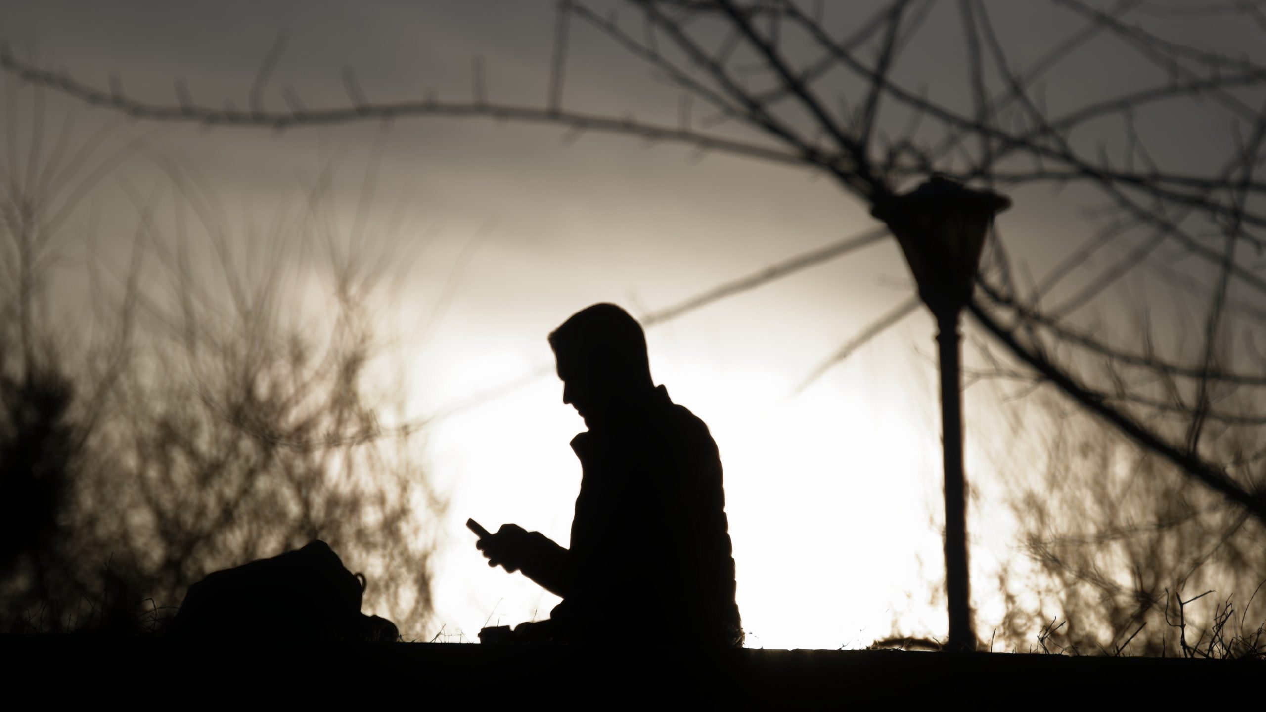Die Silhouette einer Person, die im Freien sitzt und auf ihr Smartphone schaut, hebt sich von einem hellen Himmel ab. Im Hintergrund sind kahle Baumzweige und ein Laternenpfahl zu sehen, die der Szene eine stimmungsvolle, kontemplative Atmosphäre verleihen. In der Nähe liegt eine Tasche.